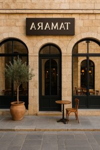 חזית מסעדת TAMARA בירושלים – שלוש קשתות אבן ירושלמית עם חלונות ודלת שחורים, שלט מודרני בצבע כהה עם כיתוב TAMARA, עציץ חרס עם עץ זית ושולחן קטן עם כיסא עץ חום בכניסה.